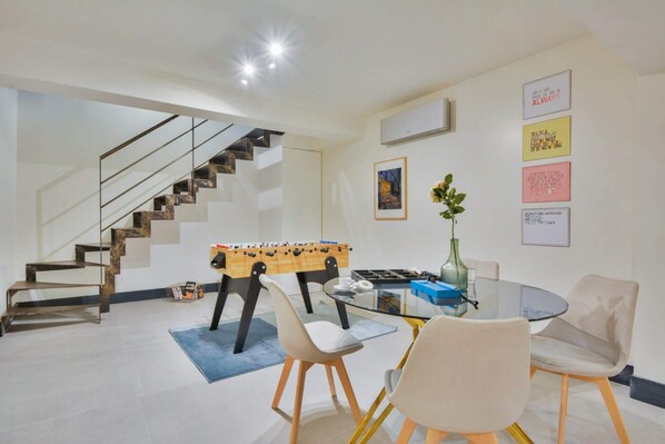 Un salon moderne avec un baby-foot, une table basse en verre et un canapé. On aperçoit un escalier au fond.