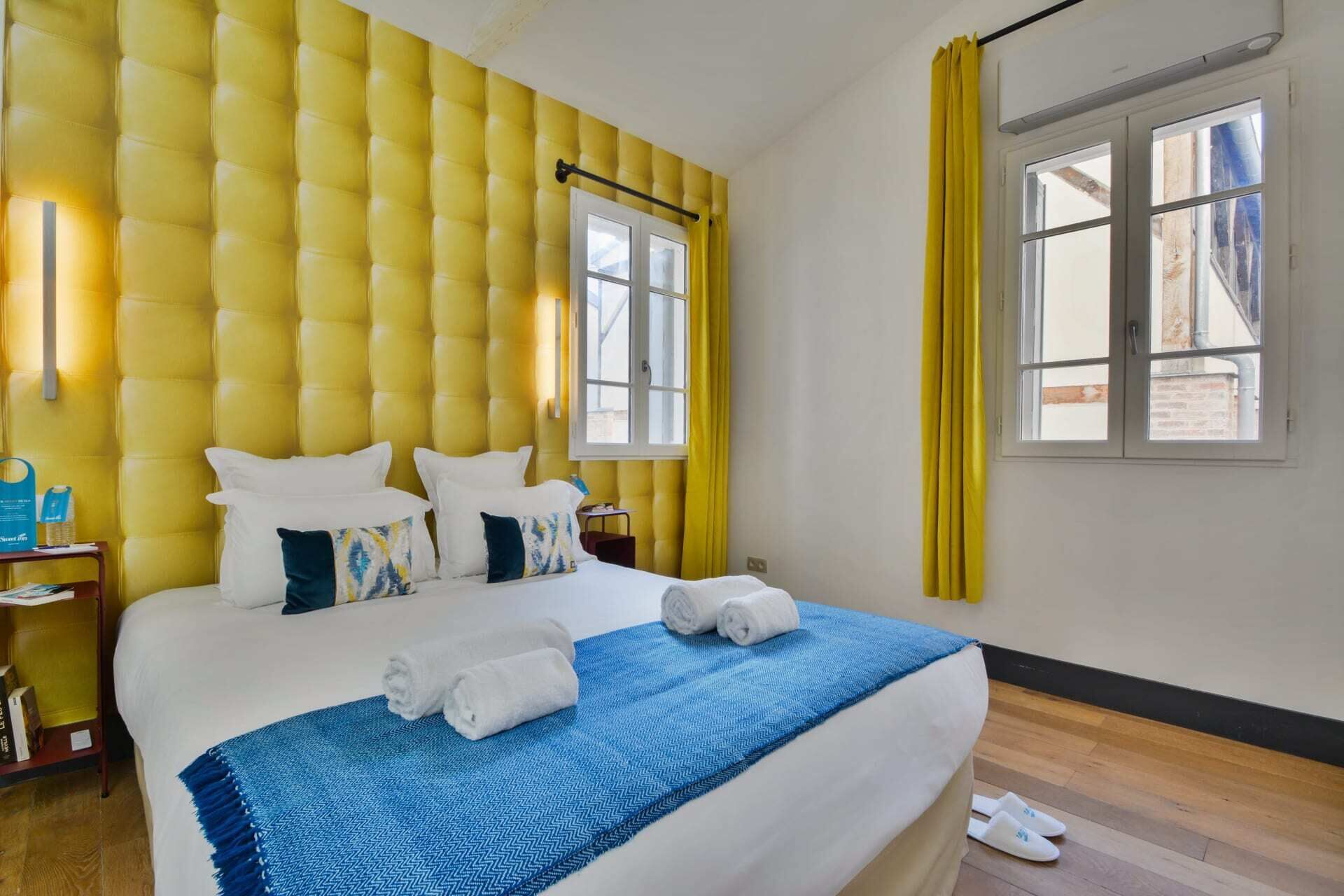 A hotel room with a tufted yellow headboard, a large bed with white linens, two windows with yellow curtains, and a wooden floor.