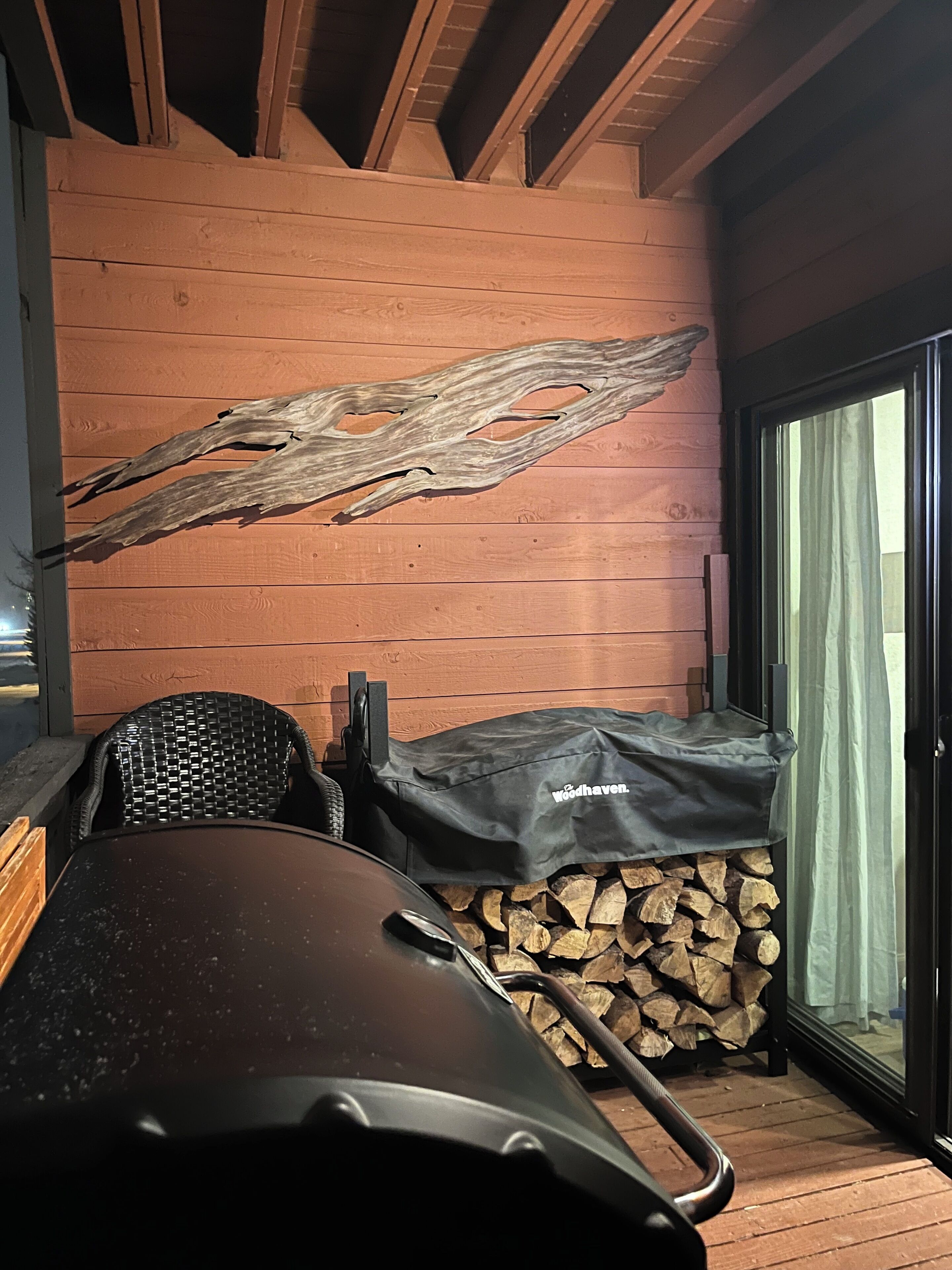 Un espacio de estar al aire libre techado, con una parrilla, leña apilada y un muro de madera con un trozo de madera a la deriva singular.