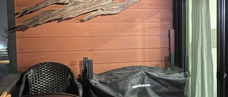 Un espacio de estar al aire libre techado, con una parrilla, leña apilada y un muro de madera con un trozo de madera a la deriva singular.