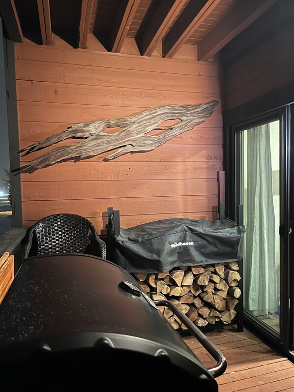 A covered outdoor seating area with a grill, a stack of firewood, and a wooden wall with a unique piece of driftwood.
