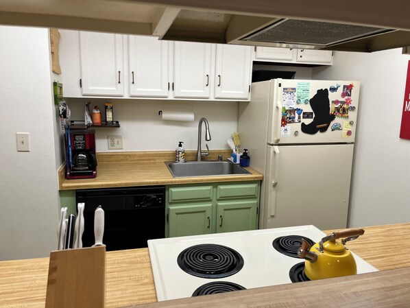 A compact kitchen with white cabinets, a stainless steel sink, a coffee maker, a dishwasher, and a refrigerator with magnets.