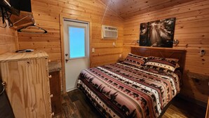 A wooden cabin bedroom with a bed, a door, a window, a ceiling fan, and a painting on the wall. - Christopher Creek Cabins - Cabin 5 (Payson)