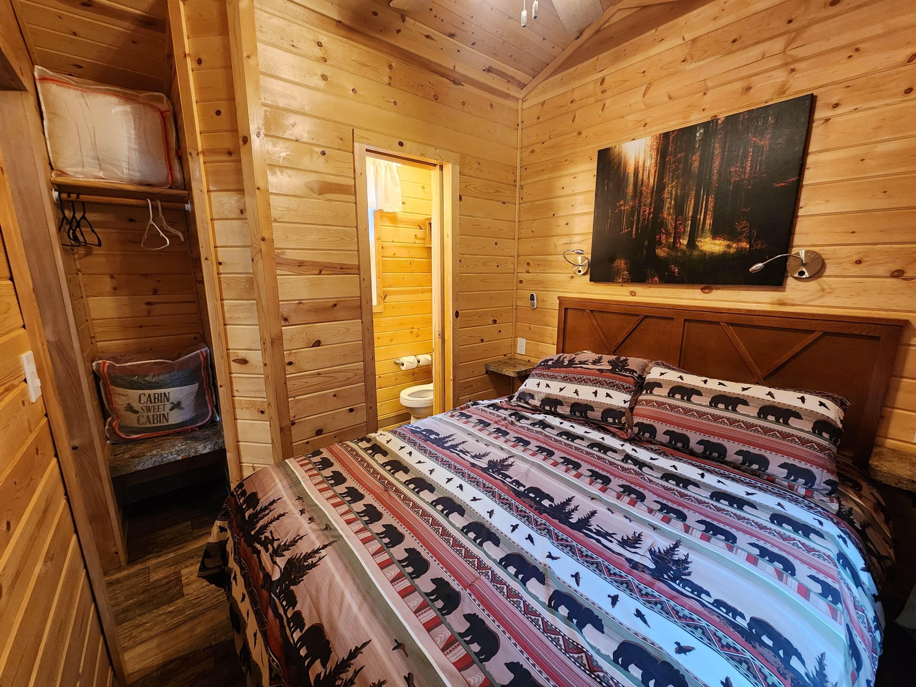 A cozy cabin-style bedroom with a wooden interior, a bed with patterned bedding, a painting on the wall, and a small bathroom visible through an open door.