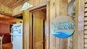 A wooden interior with a refrigerator, a door with a sign reading "Whitaker Sea La Vie," and a woven basket on the wall. - Whitaker Sea La Vie | Soundfront Property, Great Views! (Topsail Beach)