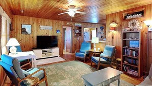 A wooden cabin interior with a television, blue chairs, a small table, and a bookshelf. - Whitaker Sea La Vie | Soundfront Property, Great Views! (Topsail Beach)