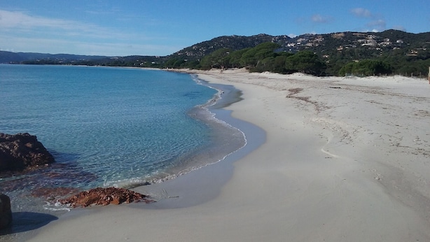 Una playa con aguas azules cristalinas, orilla de arena y un paisaje montañoso con edificios.