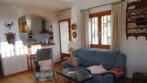 A living room with a blue sofa, a glass coffee table, and a dining area with a kitchen in the background. - Villa with private protected pool, 200 m from the beach, air conditioning  (Ametlla de Mar)