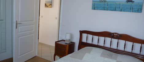 Un dormitorio con cama de madera, una mesita de noche con lámpara, una puerta que da a otra habitación y un cuadro en la pared con una escena costera.