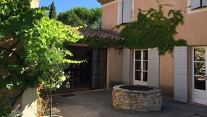 A courtyard with a stone well, a pergola covered in vines, and a building with a tiled roof and white shutters. - Provencal farmhouse from the 19th century with swimming pool, charming house (Aix-En-Provence)