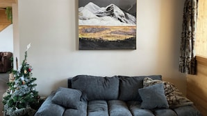 Un salon avec un canapé gris, un petit sapin de Noël et un tableau représentant un paysage de montagne accroché au mur.