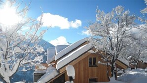 Une cabane en bois, dotée d’un toit incliné recouvert de neige, entourée d’arbres et de montagnes enneigées.
