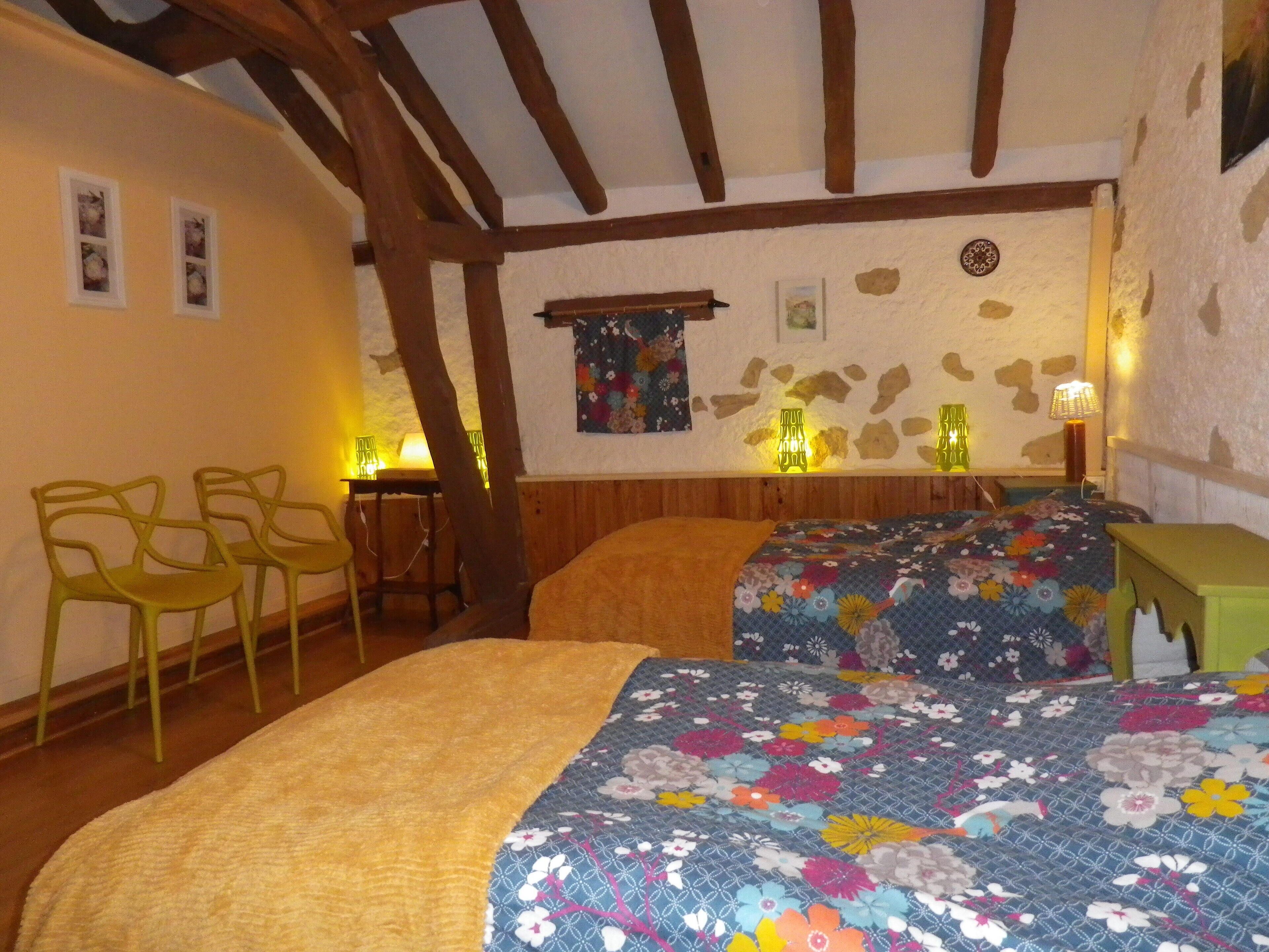 Une pièce chaleureuse avec un plafond en bois, deux lits dotés de draps à motifs floraux, une petite table, des chaises et des lampes fixées au mur.