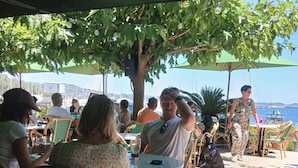 Un café al aire libre con sillas de mimbre, mesas redondas y sombrillas que dan sombra.