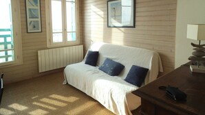 Ein Zimmer mit einer holzverkleideten Wand, einem Bett mit weißer Bettdecke und blauen Kissen, einem Holztisch mit Lampe sowie zwei mit Vorhängen versehenen Fenstern.