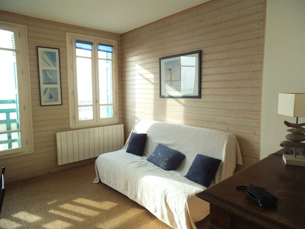 A room with a wooden paneled wall, a bed with a white cover and blue pillows, a wooden table with a lamp, and two windows with curtains.