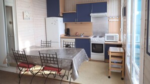 Eine kompakte Küche mit Kühlschrank, Herd, Mikrowelle und Spüle. Ein Esstisch mit Stühlen und eine kleine Holzablage sind vorhanden.
