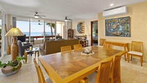 A spacious dining area with a large wooden table and chairs, a ceiling fan, and a view of the ocean. - Corner Unit in the Building That (San Miguel)
