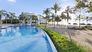 A swimming pool adjacent to a beach with palm trees and a hotel building in the background. - Corner Unit in the Building That (San Miguel)