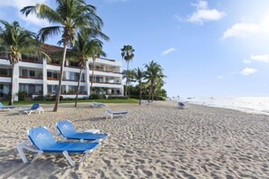 A beach with blue lounge chairs, palm trees, and a hotel building in the background. - Corner Unit in the Building That (San Miguel)