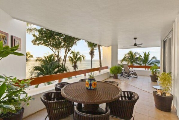 A covered outdoor area with a round table and chairs, surrounded by potted plants, overlooking a beach with palm trees. - Corner Unit in the Building That (San Miguel)