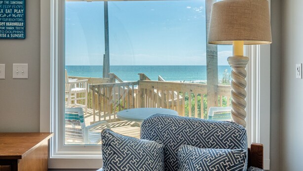 Een kamer met een groot raam met uitzicht op het strand, een houten terras en een tafel met stoelen.