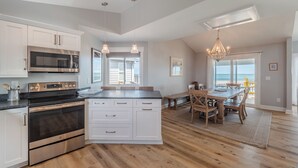 Een moderne keuken met roestvrijstalen apparatuur, een eetgedeelte met een houten tafel en stoelen, en uitzicht op de oceaan door grote ramen.