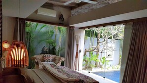 A bedroom with a bed, a nightstand, a lamp, and a view of a tropical garden through large glass doors.