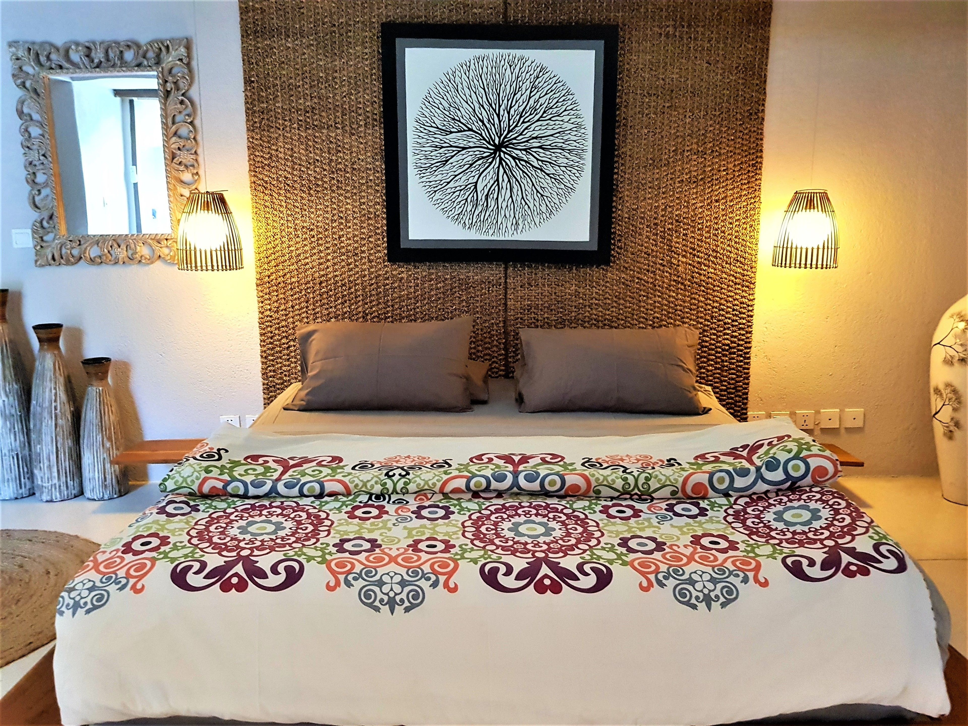 A bedroom with a patterned bedspread, two pillows, a framed artwork above the bed, and wall-mounted lights.