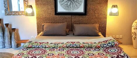A bedroom with a patterned bedspread, two pillows, a framed artwork above the bed, and wall-mounted lights.