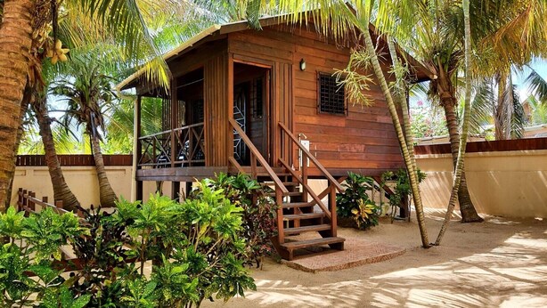 A wooden cabin with a porch, surrounded by palm trees and tropical plants. - Baby Blue Cabana (Hopkins Village)