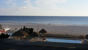 Strandudsigt med pool, liggestole og stråtækte parasoller.