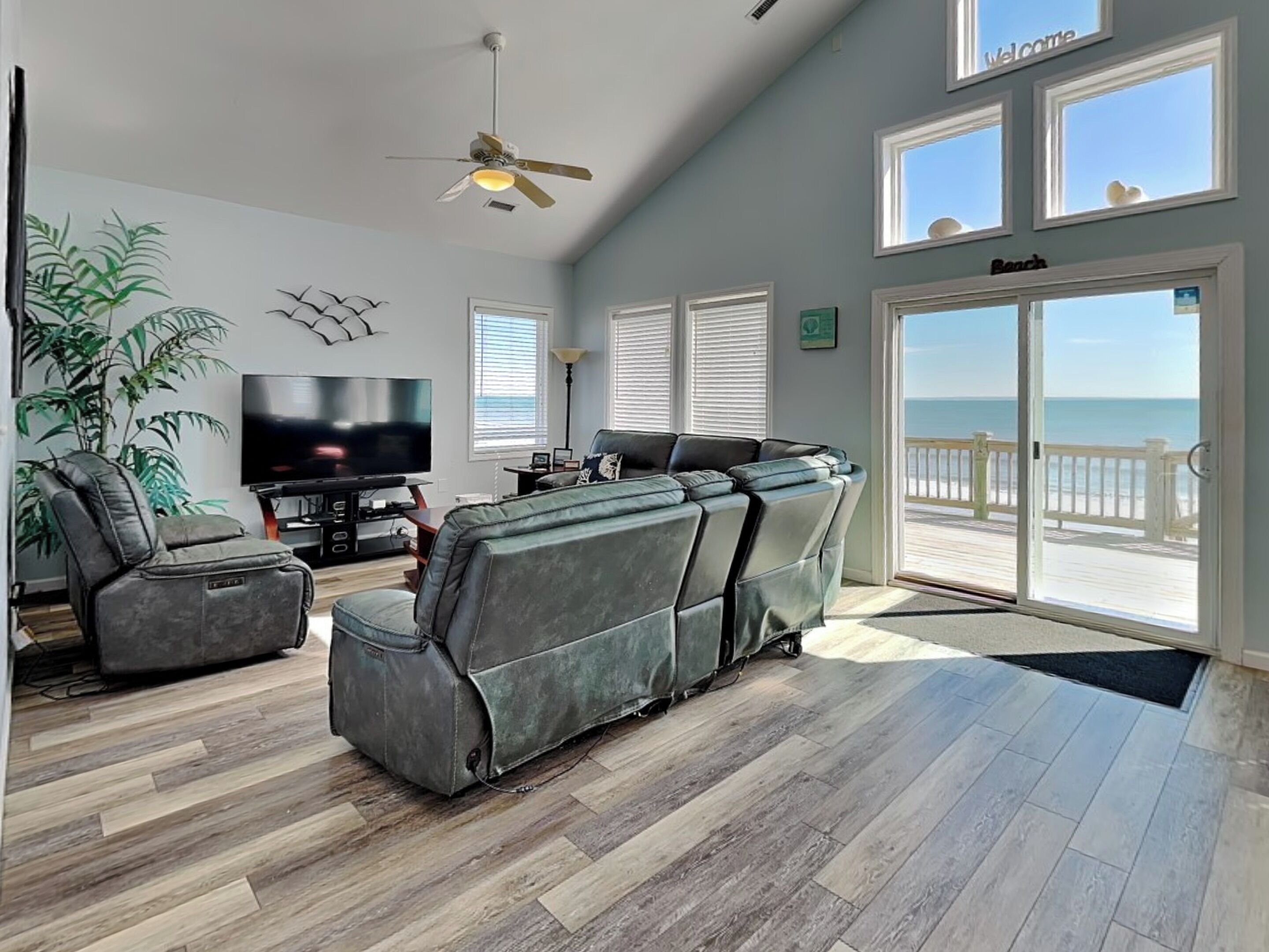 A spacious living room with a large sectional sofa, a flat-screen TV, and a ceiling fan. There are large windows offering a view of the beach.