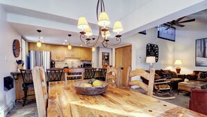 A spacious living room with a large wooden dining table, a kitchen in the background, and a ceiling fan.