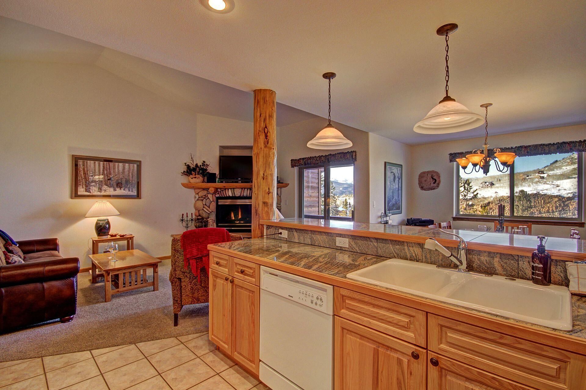 A cozy open-plan living room and kitchen area with wooden cabinetry, modern appliances, a leather sofa, a stone fireplace, large windows, and mountain views.