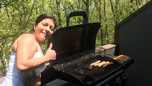 Eine Frau grillt Würstchen auf einem Grill im Freien, eine Propangasflasche steht in der Nähe.