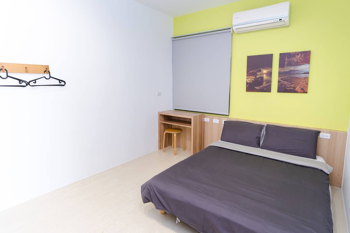 A bedroom with a bed, a desk, and a chair. There is a window with a white blind, a wall-mounted air conditioner, and two framed pictures on the wall.