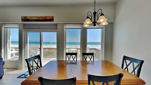 Une salle à manger avec une grande table en bois et six chaises, de grandes fenêtres donnant sur la plage et un lustre.