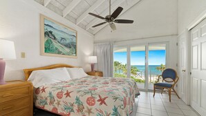 A bedroom with a bed, a ceiling fan, a painting, a chair, and a view of the ocean.