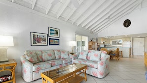 A spacious living area with a sofa, coffee table, and dining table. There are framed pictures on the wall, a kitchen in the background, and a ceiling with exposed beams.