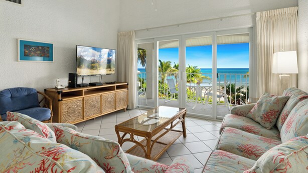 A living room with a television, a sofa, a coffee table, and a balcony with a view of the ocean.