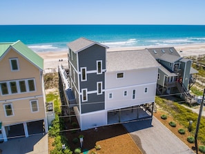 A multi-story building with a beach view, featuring a garage, a balcony, and a small outdoor area with plants.