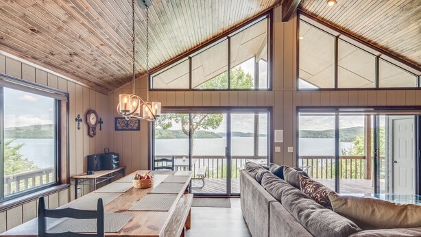 A spacious room with a wooden ceiling, large windows, a dining table, a sofa, and a view of a lake. - Spectacular 180° Beaver Lakefront Views! Steps to Shoreline+Pool Table+Fire Pit (Garfield)
