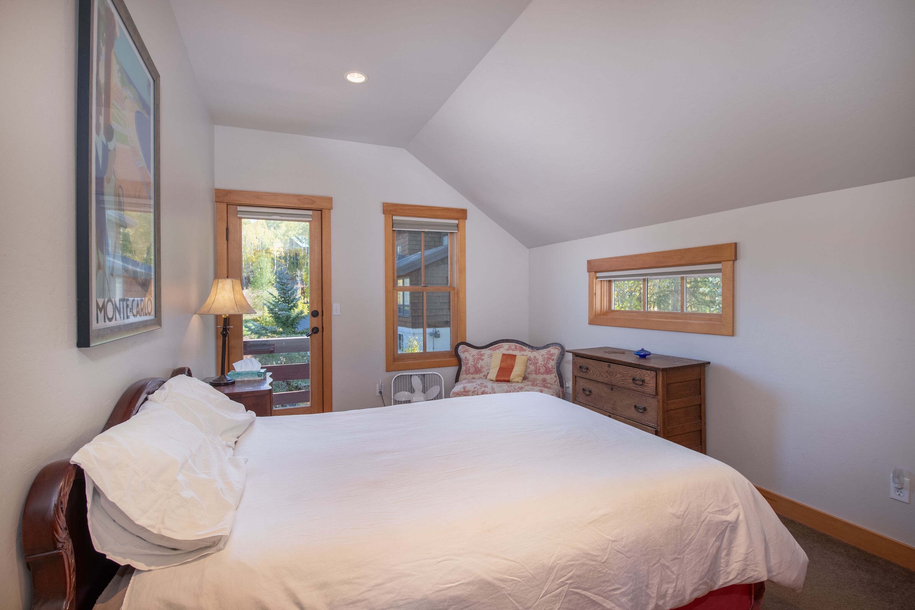 Une chambre à coucher avec un grand lit, une tête de lit en bois, une commode et deux fenêtres à cadre en bois.