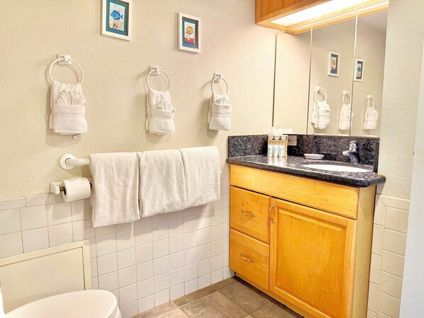 A bathroom with white tiles, a toilet, a sink with a granite countertop, and multiple towel hooks on the wall.