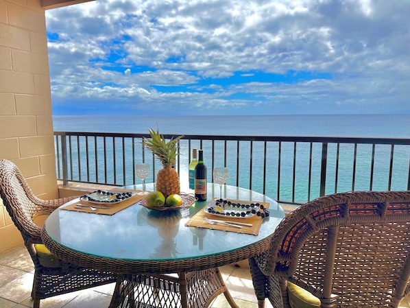 A balcony with a glass table set for two, featuring a pineapple, a bottle, and a plate of olives.