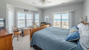 Un dormitorio con una cama grande, un escritorio y una silla. Hay ventanas con persianas, un ventilador de techo y vistas a la playa.