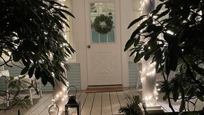 Un porxo amb una porta blanca, una corona decorativa i llums lluminoses. Hi ha plantes i fanals al porxo.