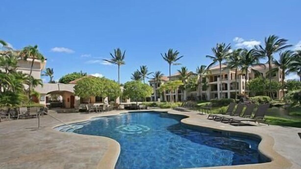 A swimming pool with a curved edge, surrounded by lounge chairs and palm trees, with buildings in the background. -  The Shores, quiet, upgraded condo, large lanai w/ garden area and lawn  (Waikoloa)