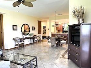 A spacious living area with a glass coffee table, wicker chairs, a dark wooden cabinet, and a flat-screen TV.